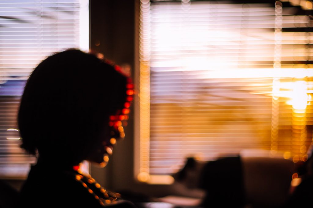 A person sitting in front of a window with blinds closed and sunlight peaking through