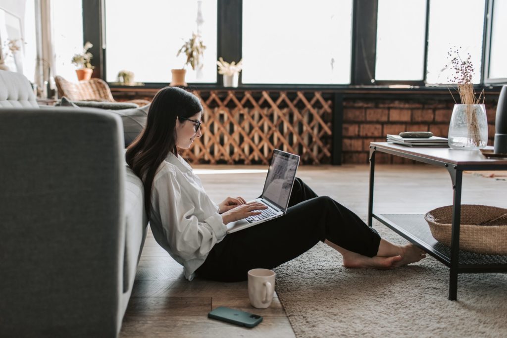Woman working from home sitting on the floor with her laptop, coffee and cell phone