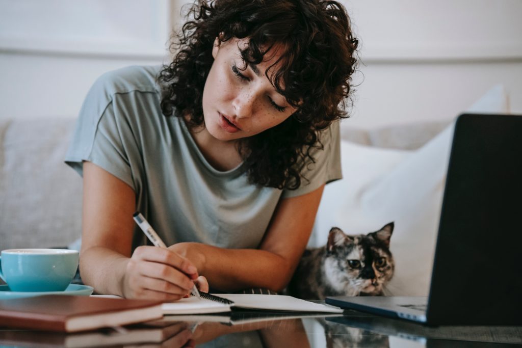 Girl writing in a notebook from the computer with her cat on the couch