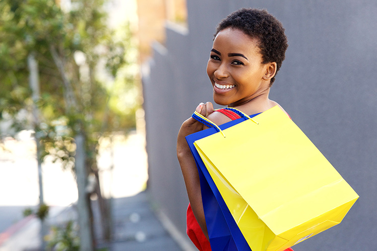 Woman looking back as she walks smiling into camera holding shopping bags over her shoulder.